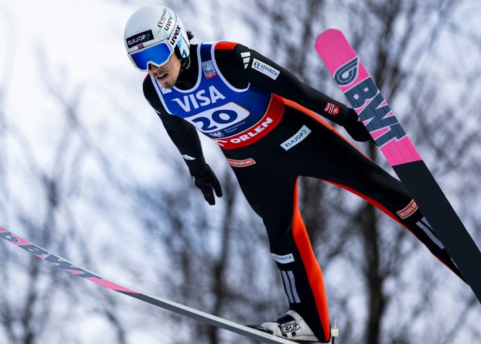 Ski jumper in mid-air wearing a black and orange suit, illustrating Olympic controversy amid hyaluronic acid manhood injections.