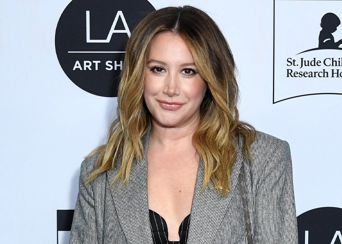 Ashley Tisdale attending a public event, wearing a grey blazer with loose wavy hair against an event backdrop.