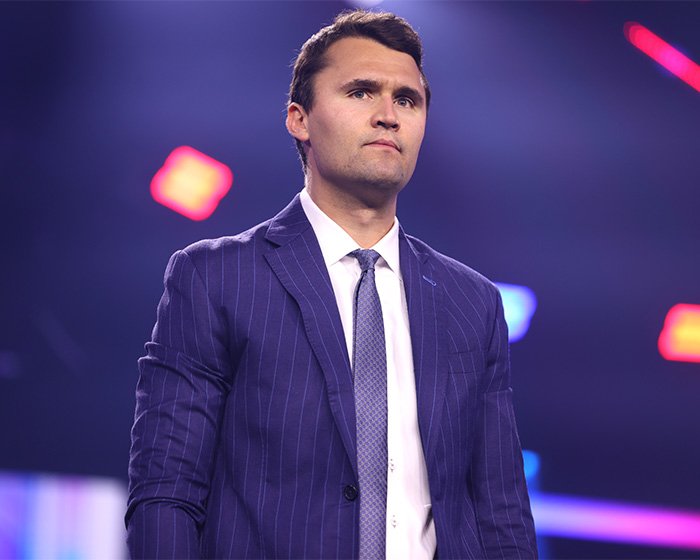 Man in a dark pinstripe suit standing on stage with colorful blurred lights in the background at an event.