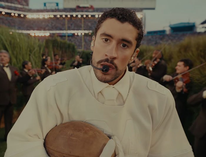 Bad Bunny wearing headset and holding football during Super Bowl performance with musicians in the background.