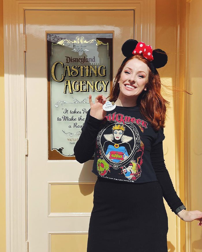 Woman wearing Evil Queen shirt and Minnie ears stands by Disneyland Casting Agency door holding a name tag, smiling.