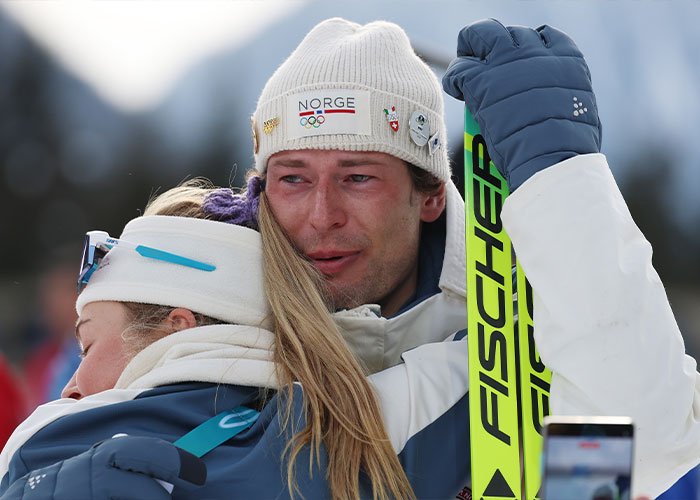 Olympic medalist in winter gear, wearing a knit cap, embracing a teammate while holding Fischer skis after a medal victory.