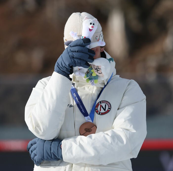 Olympian wearing white jacket and bronze medal covering face with gloves holding two plush toys outdoors.