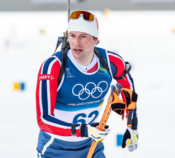 Olympian athlete competing in winter biathlon event wearing red, white, and blue with ski poles on snowy course.