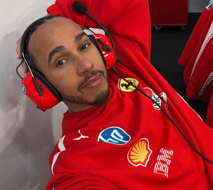 Lewis Hamilton wearing red racing gear and headphones, showcasing body language expert analysis of tightly controlled moves.
