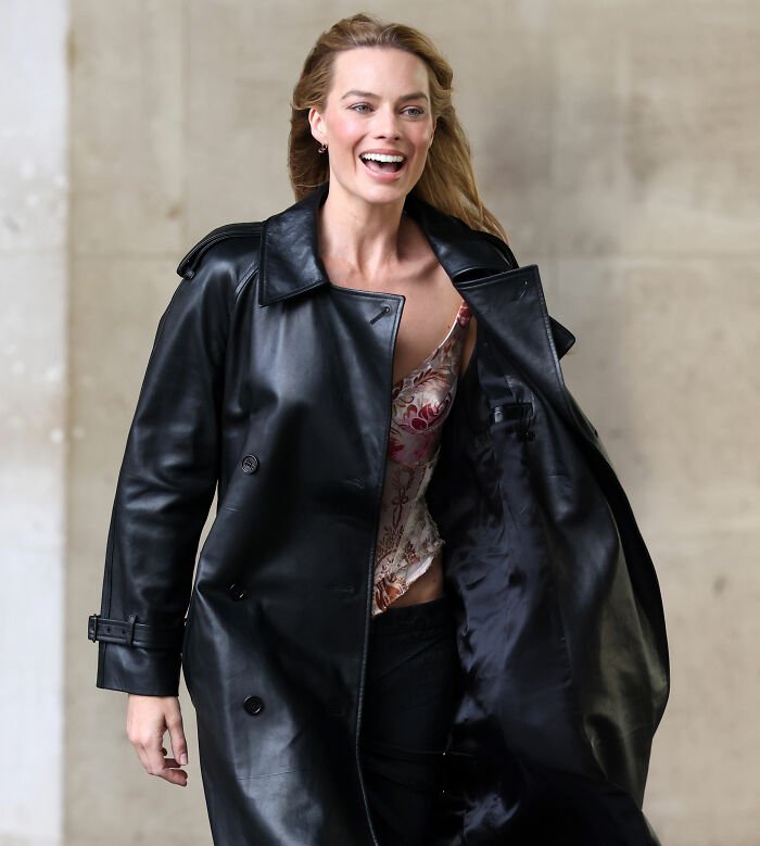 Margot Robbie wearing a black leather coat, smiling and walking outdoors, related to gift male actor gave her controversy.