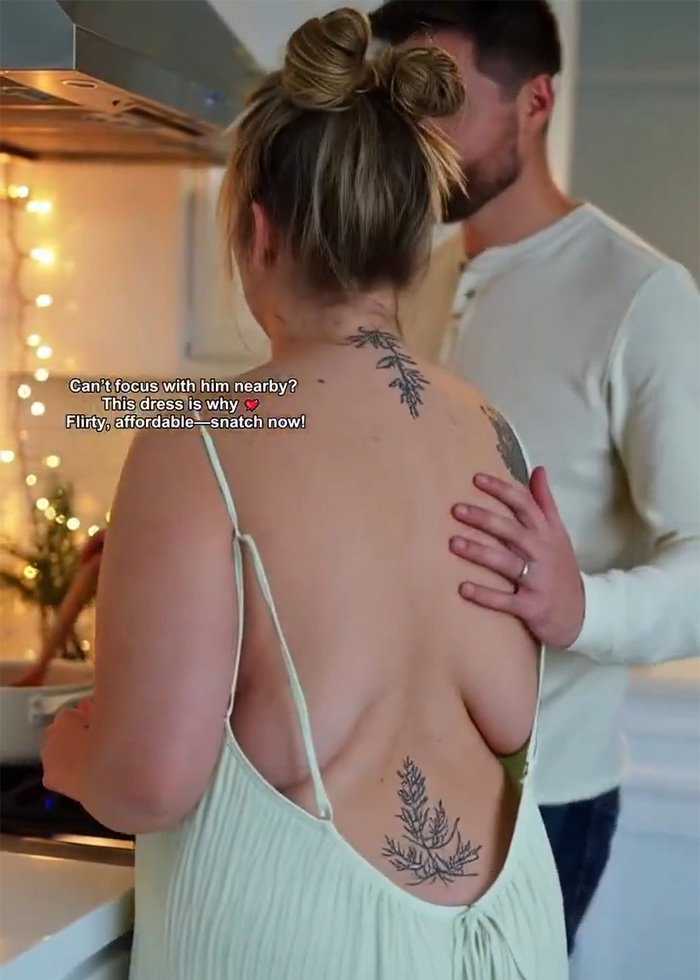 Woman wearing a low-cut dress with floral back tattoos, standing in a kitchen with a man touching her shoulder.