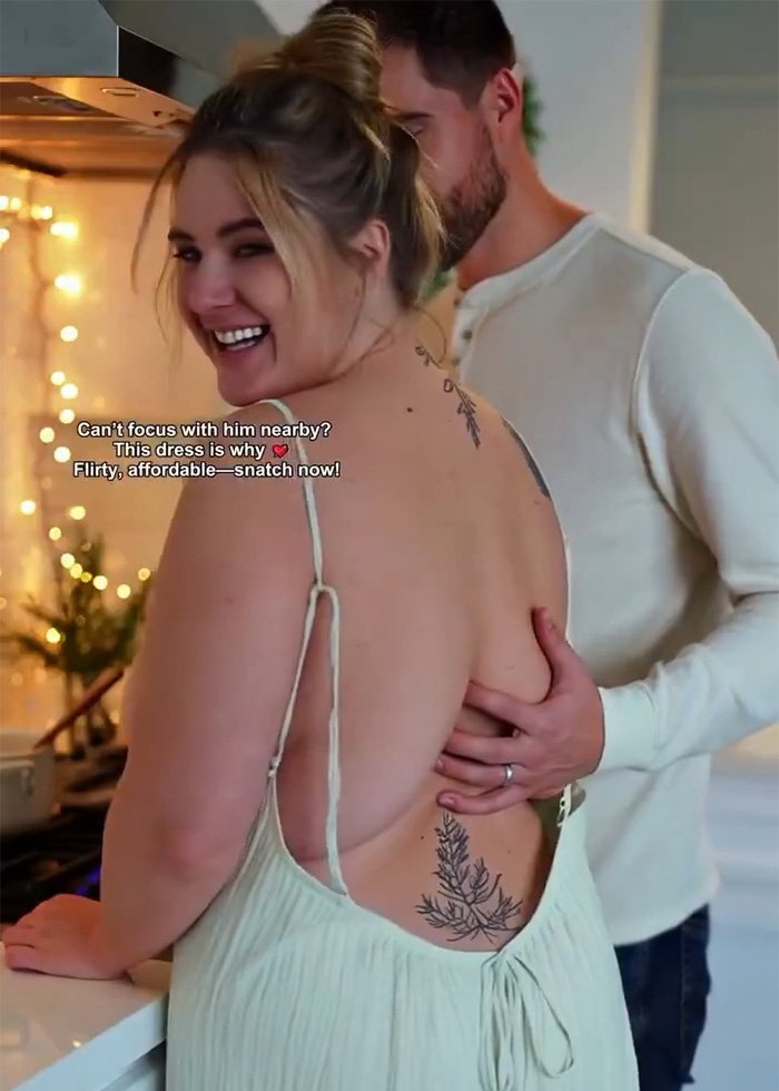 Woman smiling in a low-cut dress with tattooed back while a man stands close behind her in a cozy kitchen setting.