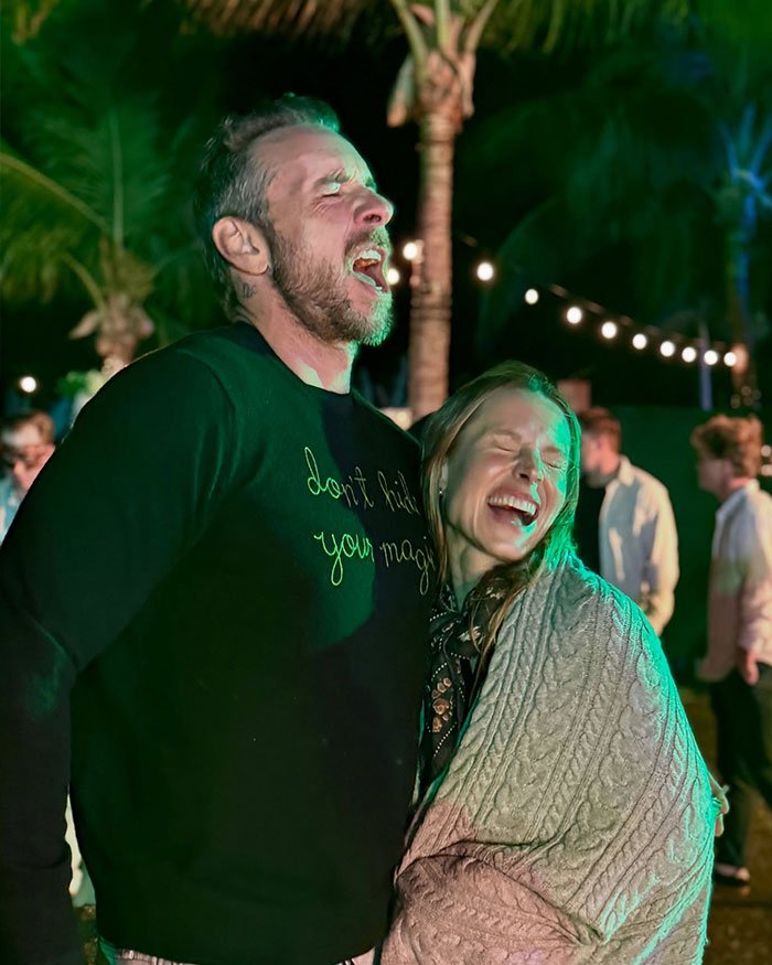 Dax Shepard laughing and embracing Kristen Bell outdoors at night, sharing a candid moment under string lights and palm trees.