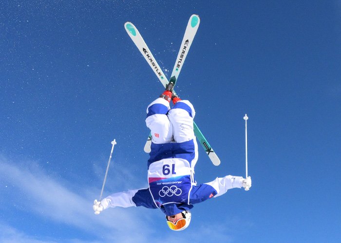 Trans Olympian performing an aerial skiing trick in Olympic gear under a clear blue sky during competition.