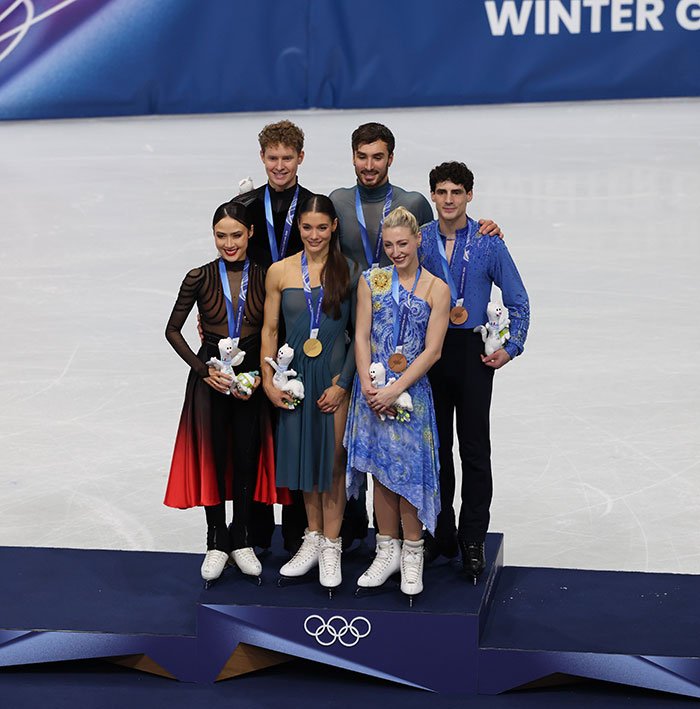 French skater on Olympic podium with medal, holding mascot, amid cheating scandal and suspicious win discussions.