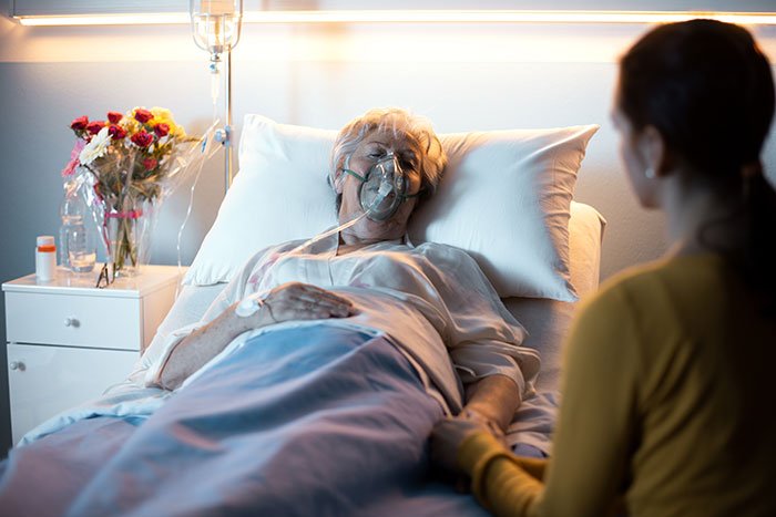 Elderly woman in hospital bed with oxygen mask, cared for by a younger woman holding her hand in a dimly lit room.