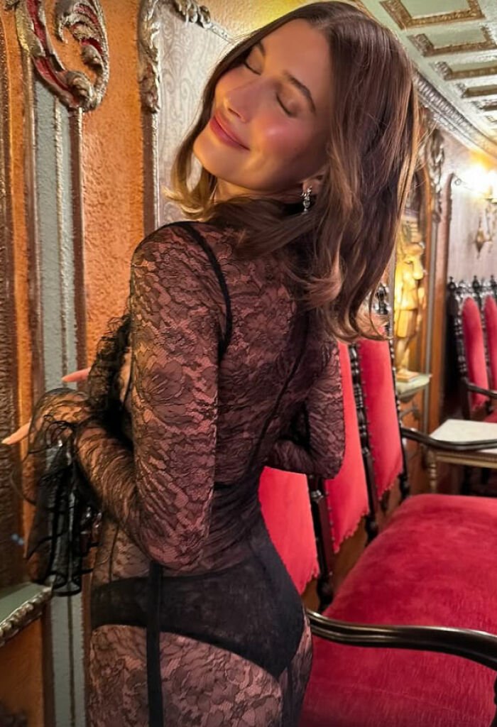 Hailey Bieber wearing black lace lingerie outfit posing inside an ornate room with red velvet chairs and wooden decor.
