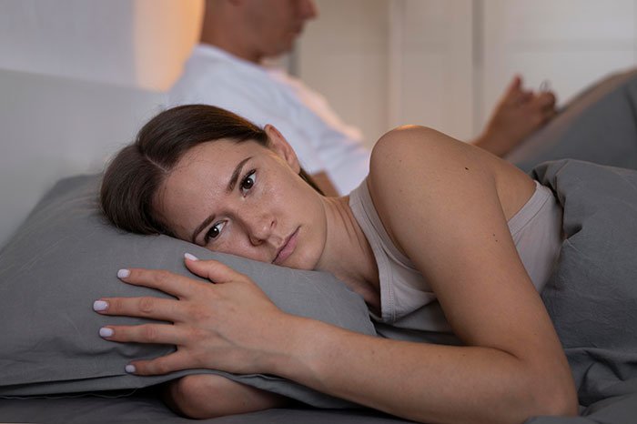 Married man texting woman he loved 15 years ago as wife lies awake in bed looking upset and distant.