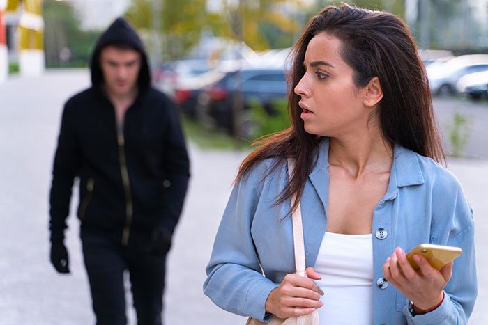 Woman holding phone looks back worriedly while a man in a black hoodie follows her, illustrating safety tips for women.