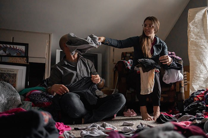 Man feels embarrassed by girlfriend’s style as they sit among scattered clothes in a dimly lit room.