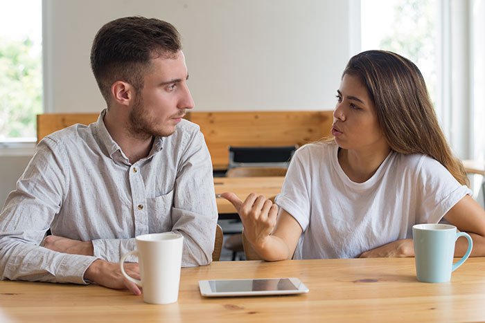 Man and woman having serious conversation at table with coffee mugs, man looking embarrassed by girlfriend’s style.