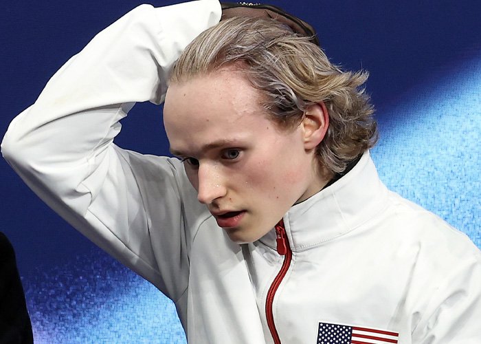 Ilia Malinin in a white jacket with an American flag patch, showing a tense expression after Olympic performance.