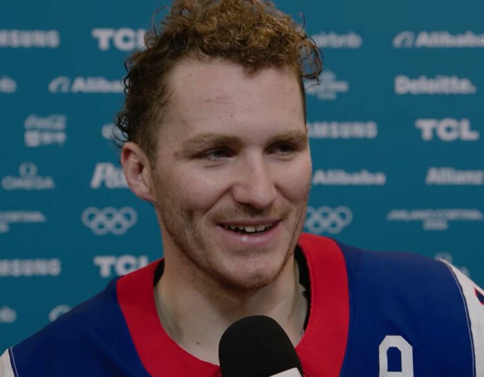 Olympic hockey player smiling during a post-game interview, wearing blue and red jersey in a media area.