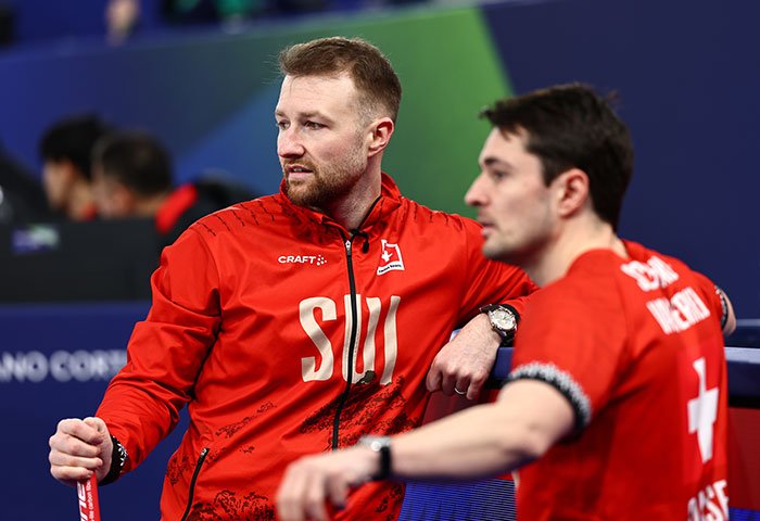 Two male athletes in red Swiss team jackets sitting at the Olympics after a major change following cheating accusations.