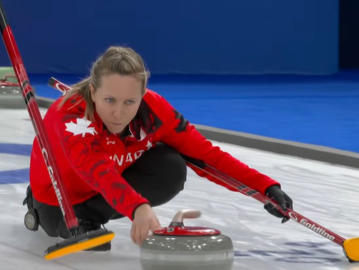 Female Canadian curler in red jacket releasing stone on ice during Olympics after cheating accusations controversy.