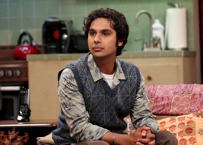 Kunal Nayyar from The Big Bang Theory seated in a living room setting, facing slightly to the side with a thoughtful expression.