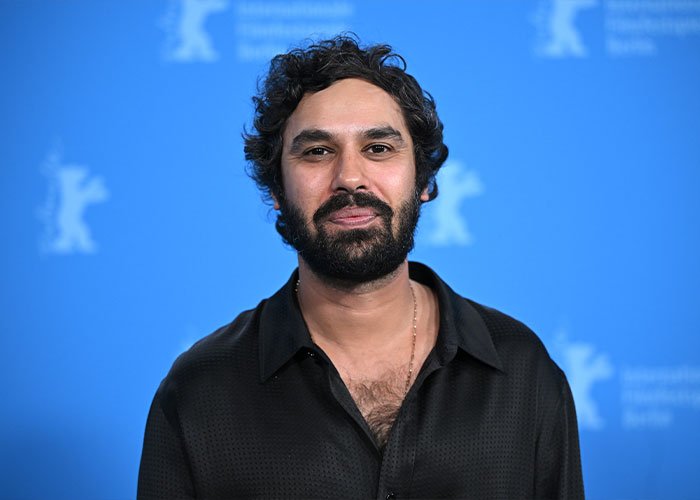 Kunal Nayyar at an event, wearing a black shirt, with a neutral expression amid a blue backdrop featuring logos.