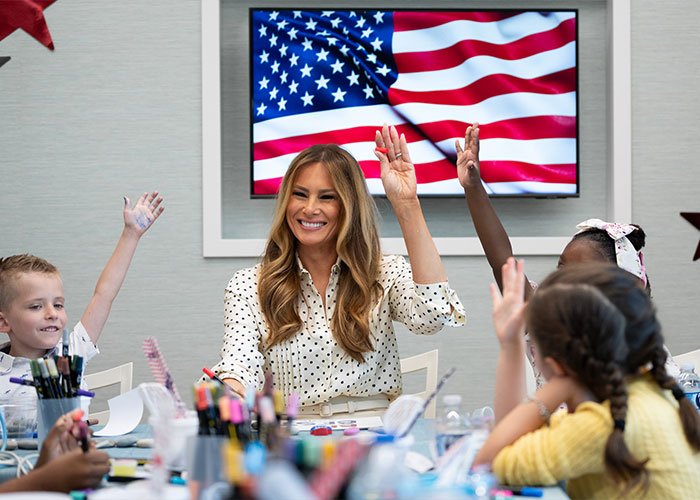 Melania Trump smiling and raising hand with children in classroom setting with American flag on screen behind.