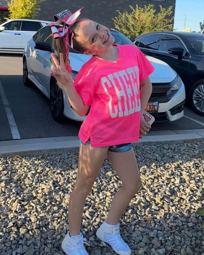 Young girl in bright pink cheer outfit posing outdoors with a peace sign, related to Utah Dance Mom’s mean texts and parent clash.