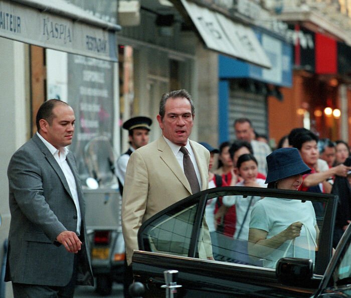 Tommy Lee Jones arriving at a city street scene with people and officials near a black car in a busy urban area.