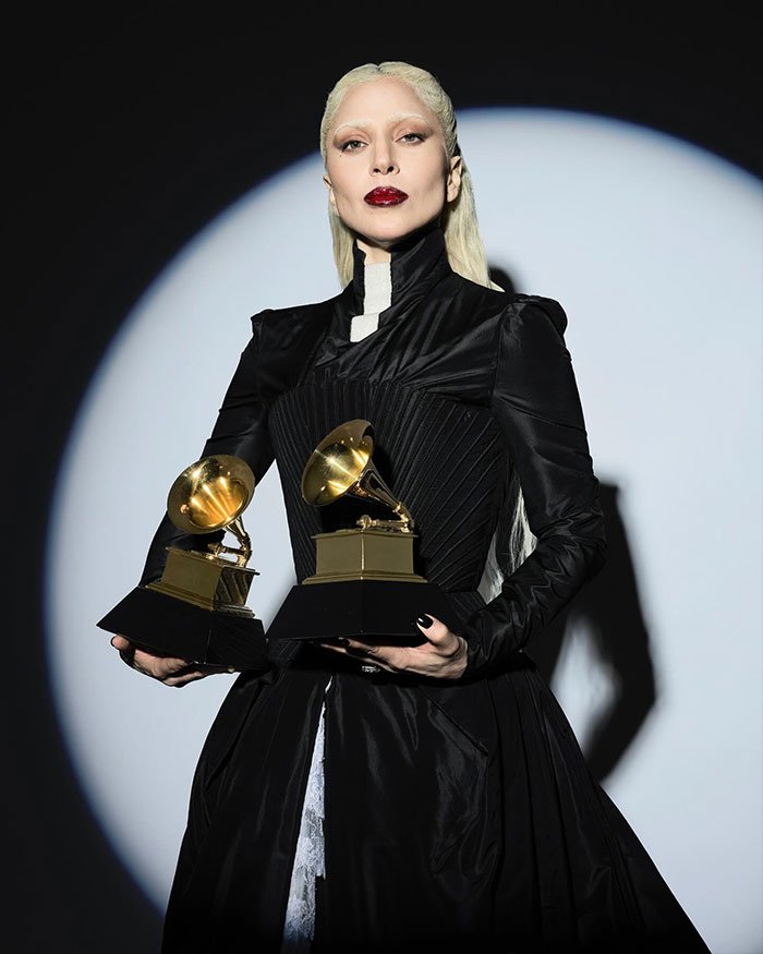 Lady Gaga with a scary look and disturbed mannerisms holding two Grammy awards on stage in dramatic lighting.