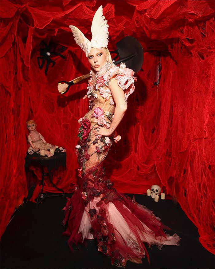 Lady Gaga in a scary floral gown and white bunny ears, holding a shovel in a red room with creepy decorations.