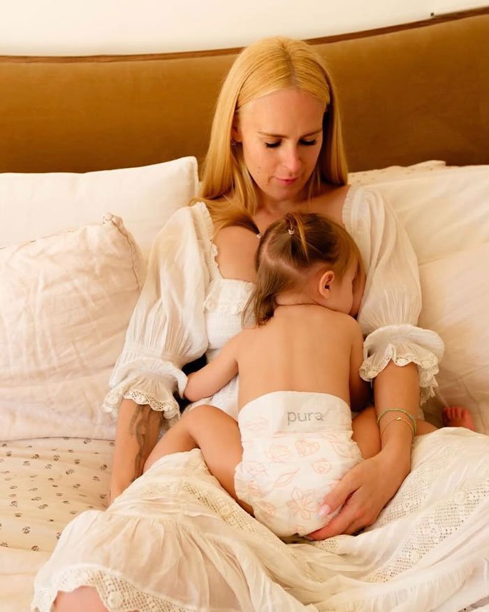 Rumer Willis sitting on a bed, wearing a white dress, holding a toddler in a diaper in a cozy home setting.