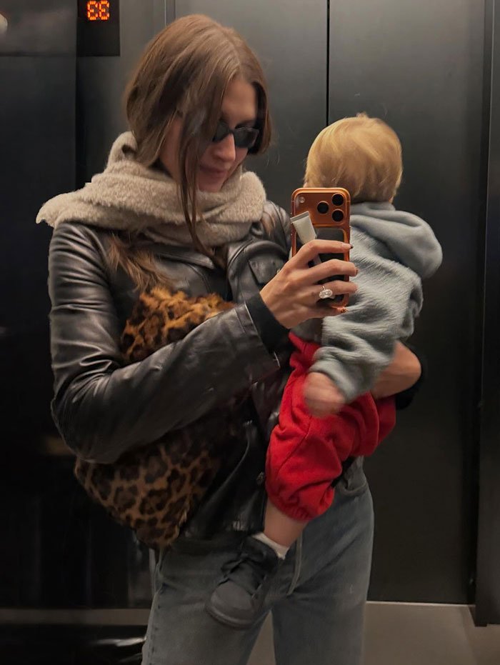 Hailey Bieber in sunglasses and leather jacket holding toddler son, capturing a mirror selfie in an elevator.