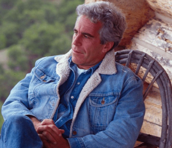 Man in a denim jacket sitting outdoors against a wooden wall, linked to Erika Kirk's alleged Epstein conspiracy theories.