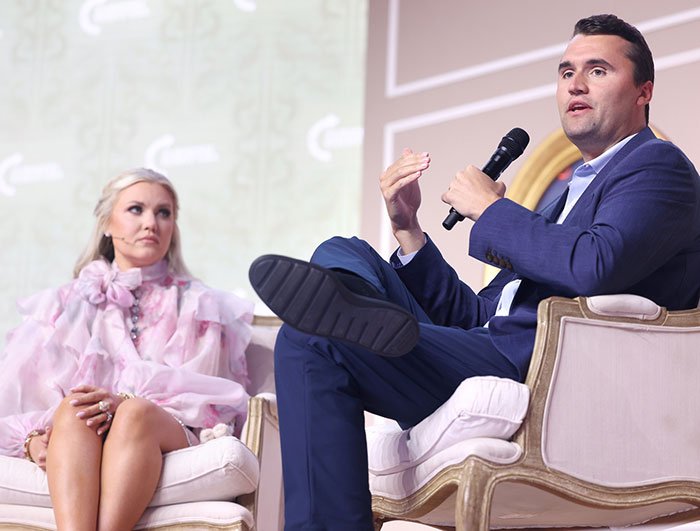Man in blue suit speaking into microphone while seated next to woman in pink dress amid Epstein link conspiracy discussion.