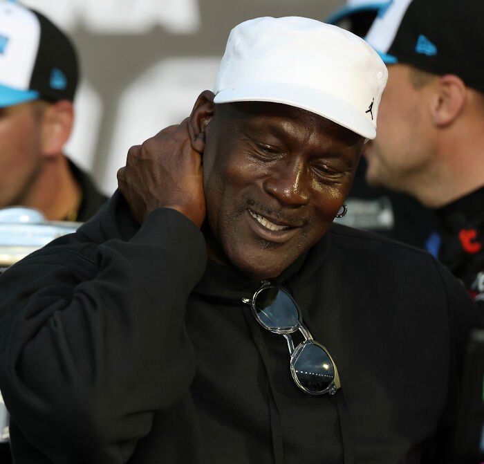Michael Jordan wearing a white cap and black shirt, smiling during a NASCAR driver controversy event.