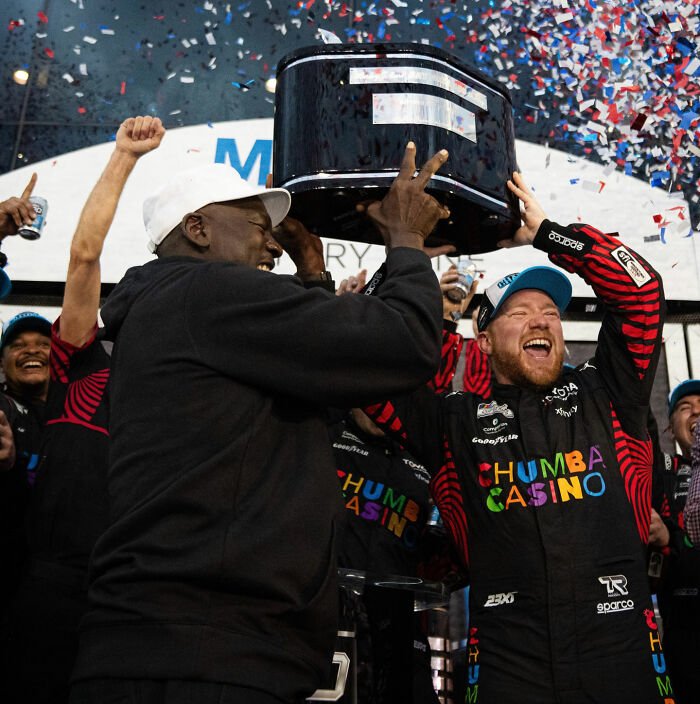 NASCAR driver celebrating victory with team and trophy amid confetti after Michael Jordan interaction controversy.