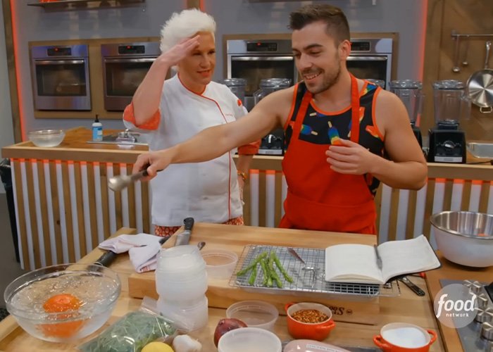 Anne Burrell cooking in a kitchen, smiling and interacting with a young man while preparing food ingredients.