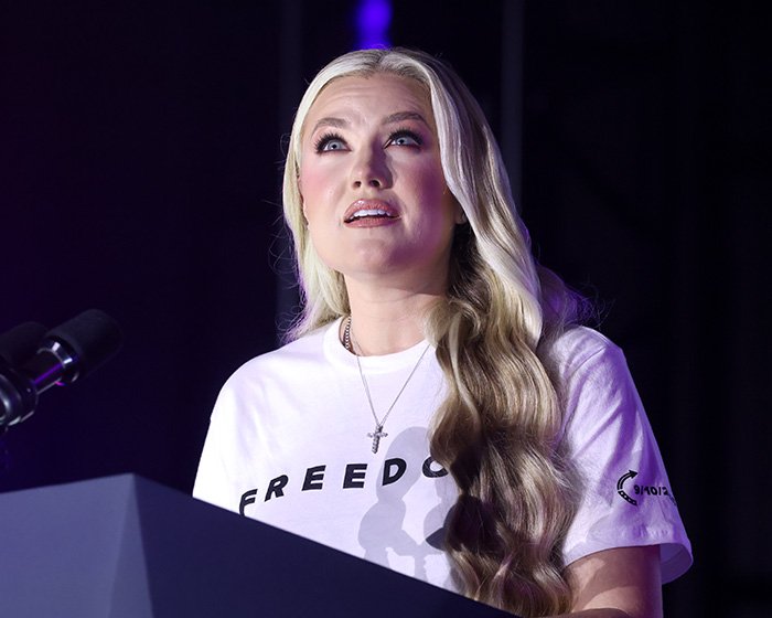 Erika Kirk speaking at a podium wearing a white freedom t-shirt with long blonde hair under purple lighting.