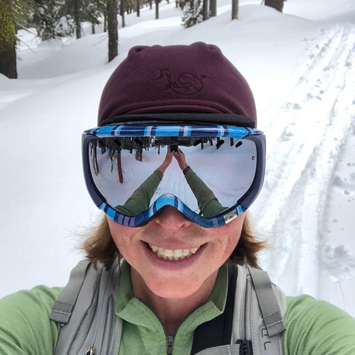Woman wearing ski goggles and winter hat smiling in snowy forest, related to mothers tragic incident outdoors.