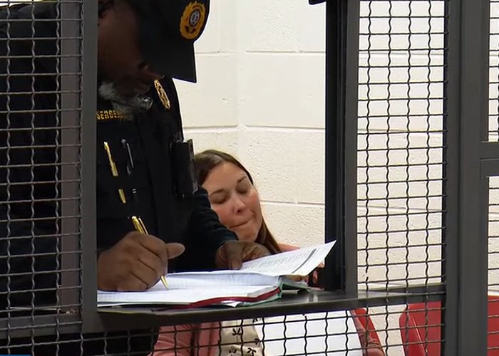 Police officer processing teacher jailed over alleged racist attack inside a detention area with woman sitting in the background.