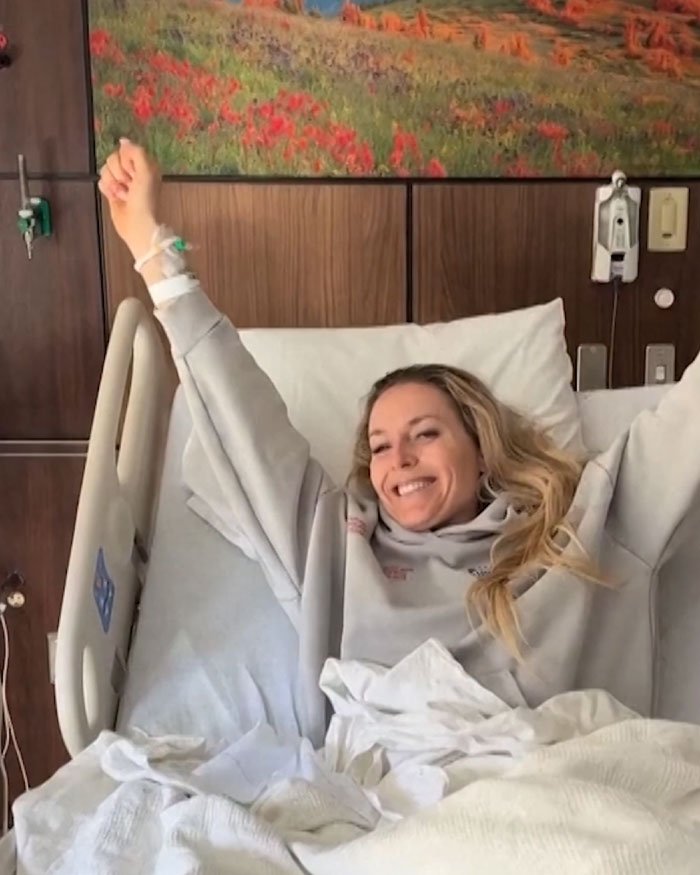 Lindsey Vonn smiling in hospital bed following fifth surgery on broken leg, raising her arms in a hopeful gesture