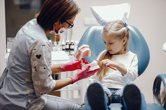 Dentist in gloves showing a child how to brush teeth using a dental model, addressing throat pain concerns.
