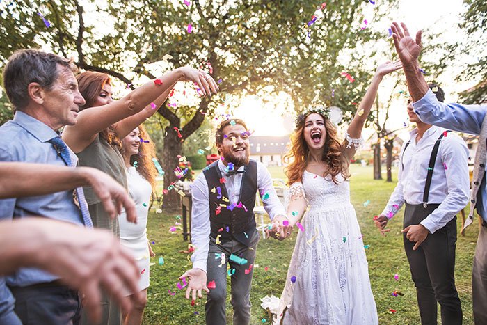 Bride laughing joyfully with friends throwing confetti outdoors, highlighting maid of honor duties at a wedding celebration.