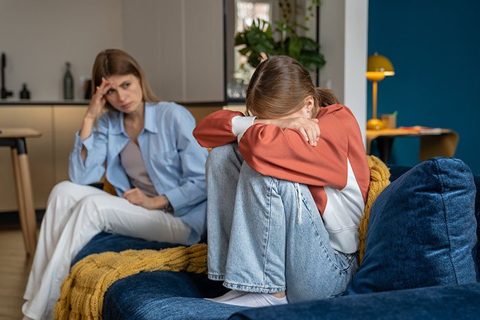 Teen sitting with head down on couch while adult woman watches worried, depicting regret and family trouble with CPS.