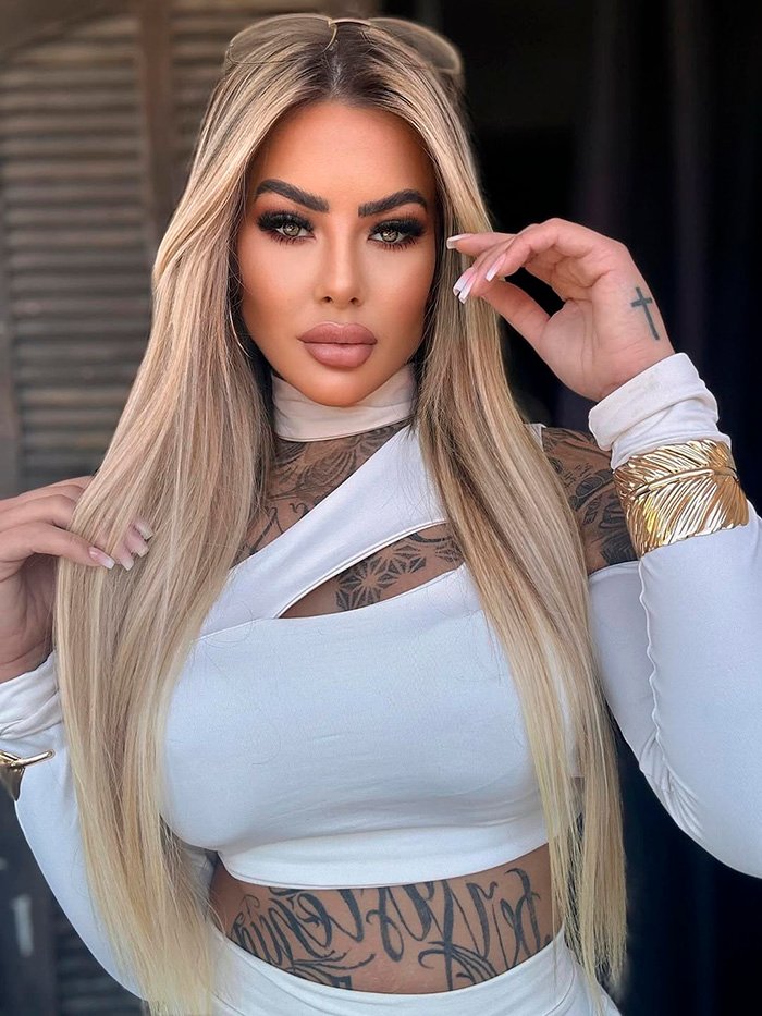 Glamorous influencer with long blonde hair and tattoos posing in a white outfit, highlighting major plastic surgery results.