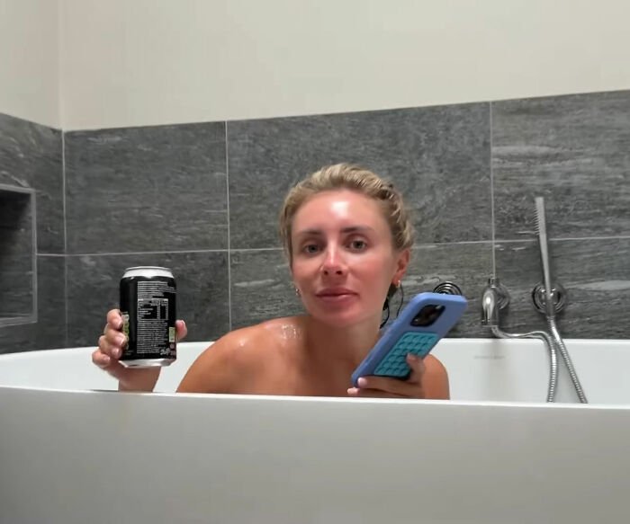 Woman in bathtub holding phone and drink, relating to Bonnie Blue pregnancy after 400-men challenge announcement.