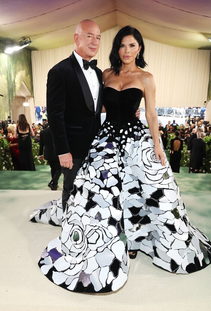 Couple posing at a fashion event, woman wearing a floral black and white ball gown, man in a black tuxedo, Met Gala dress code controversy.