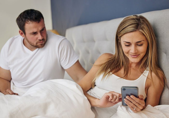 Husband looking concerned while wife smiles at phone in bed, hinting at wife&rsquo;s secret exposed after childbirth.
